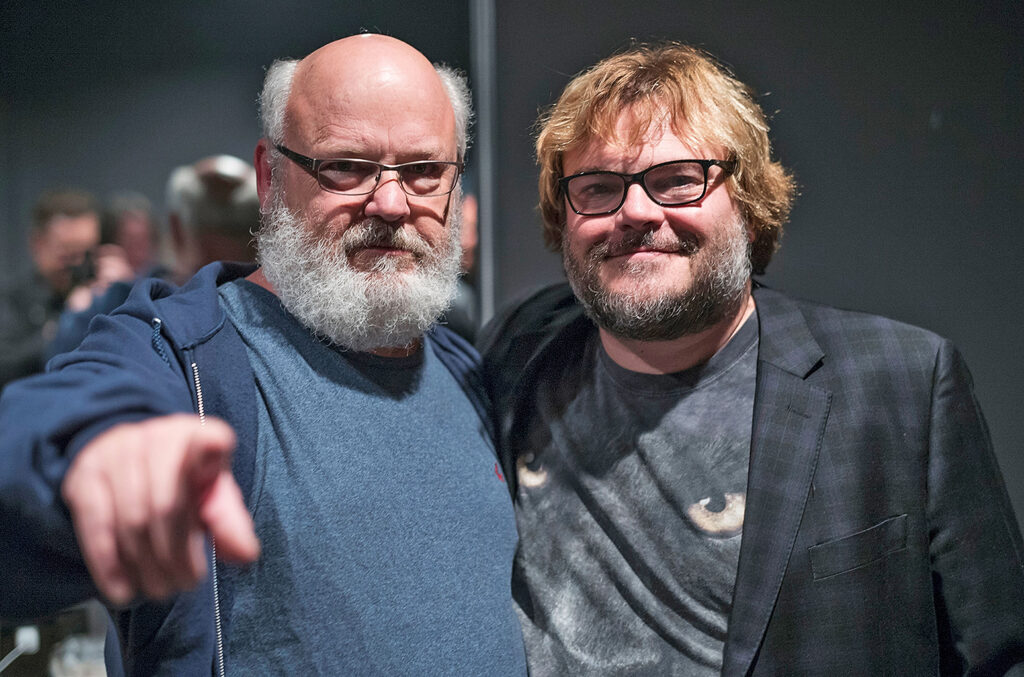 Kyle Gass and Jack Black of Tenacious D smiling for the camera. Kyle is wearing a light blue shirt with a blue hoodie, and Jack is wearing a black cat t-shirt with jacket overcoat.