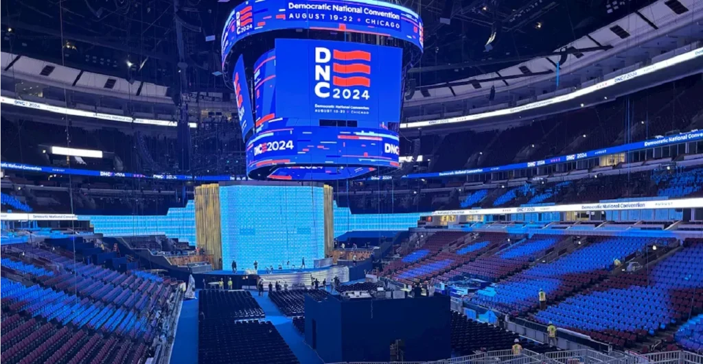 An image of the Democratic National Convention for 2024, an arena, empty, bathed in blue light with a jumbotron image of the DNC logo, the white letters DNC 2024, and a 3 striped red flag.