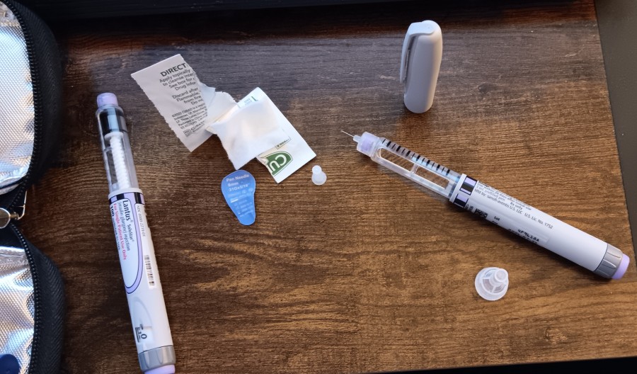 An image of several insulin pen needles, an alcohol wipe, and a pen needle on a wooden desk as background, next to an open soft case that carries insulin.