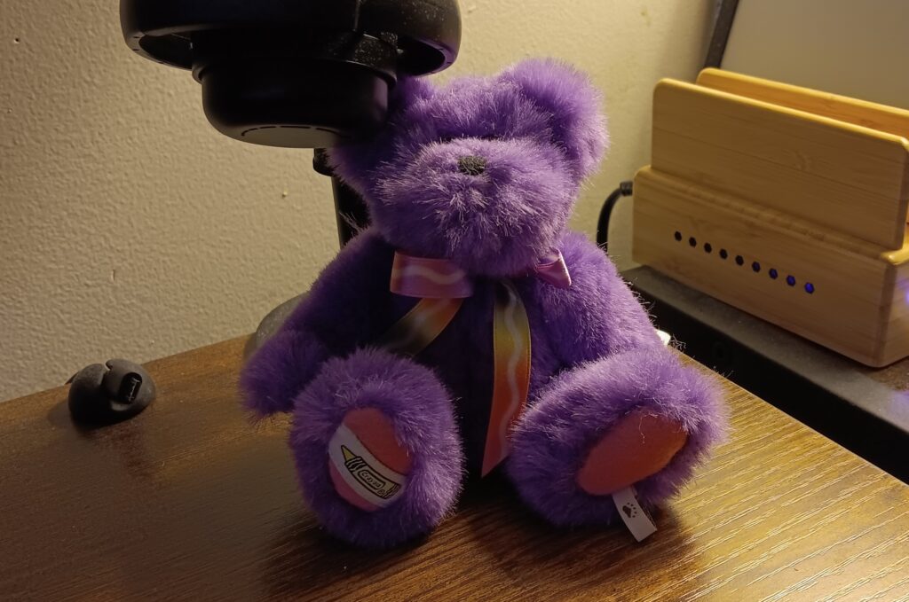 An image of a small, fuzzy purple bear plush with a tag marked 'Crayola' on the bottom right foot.