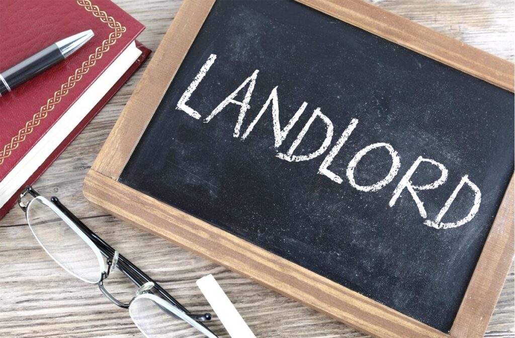 The image of a chalkboard with the word "landlord" scrawled across it. At the bottom of the image is a pair of glasses, a piece of chalk, and to the left the edge of a red book and a black pen.