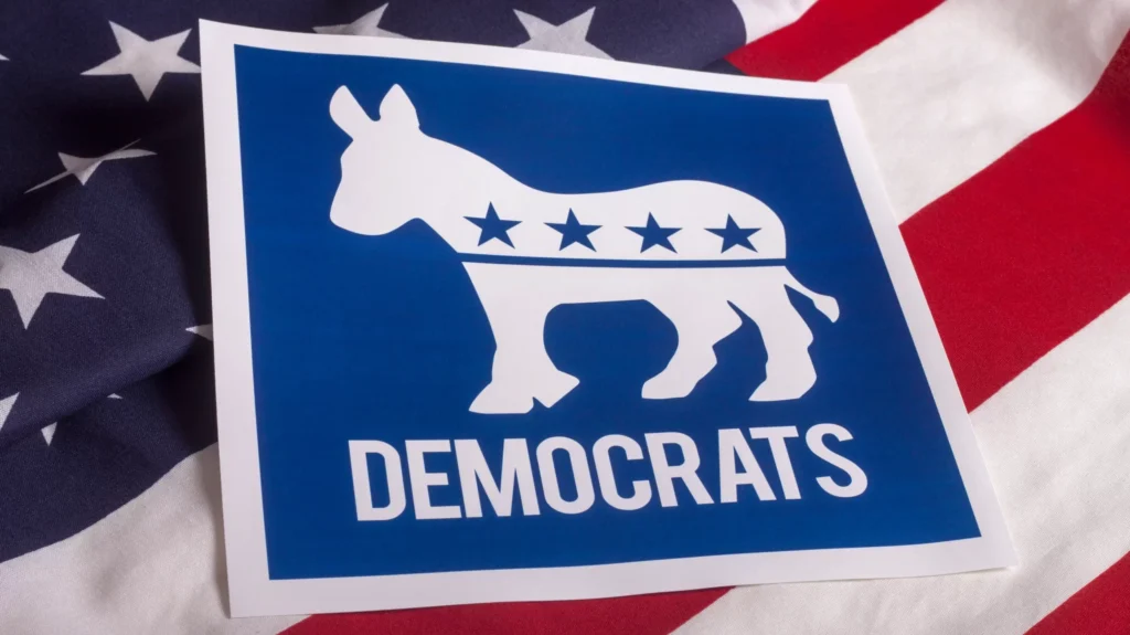 An image of the US flag in the background, and on top of it is a sign, white outline with blue filling in the negative spaces, it has the white outline of a donkey with the four stars representing the Democratic party, and it reads "Democrats."