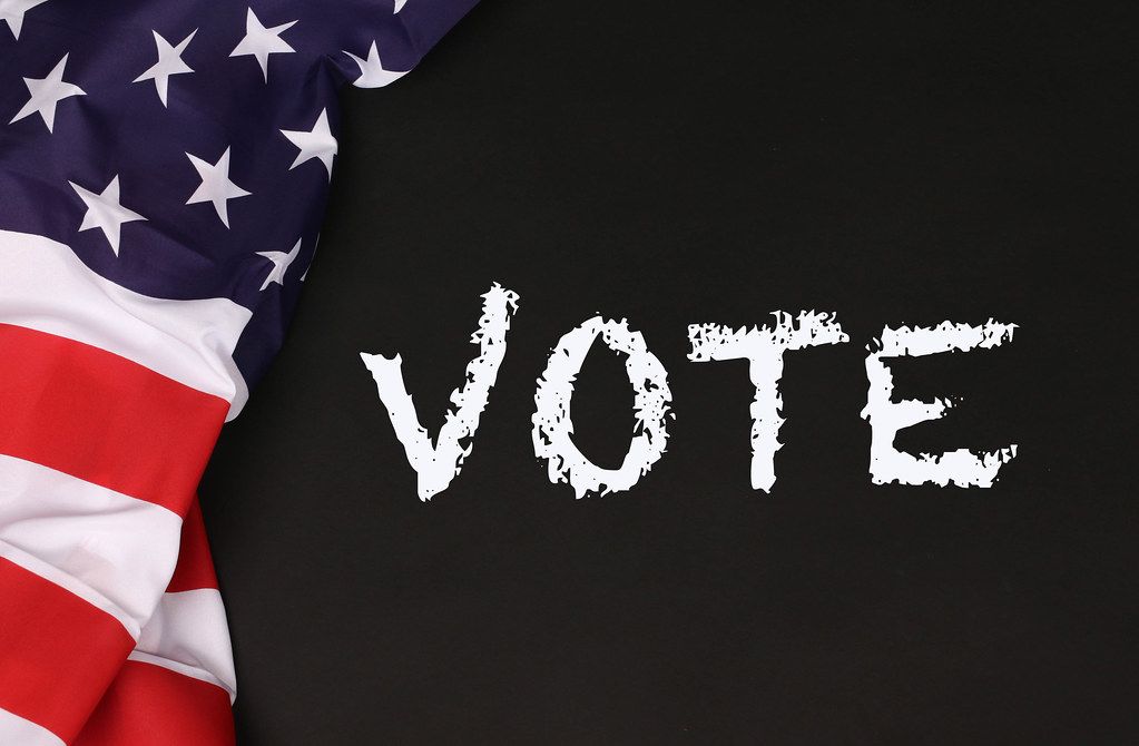 An image of a folded flag on the left hand side of a chalkboard background, with the word "vote" written in a chalk like font.