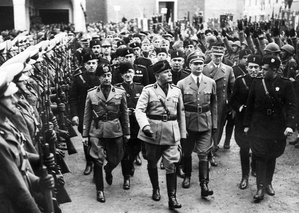 An image of Italian fascist and dictator Benito Mussolini walking with his generals along a dirt road as a phalanx of soldiers line the sides of the road, standing at attention.