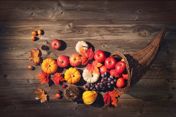 An image of a cornucopia, also known as a horn of plenty. The image shows a background of wooden slats, and sitting on this is a woven horn shaped basket, in which there are red applies, yellow squash, and purple grapes spilling out, along with a few autumn leaves.