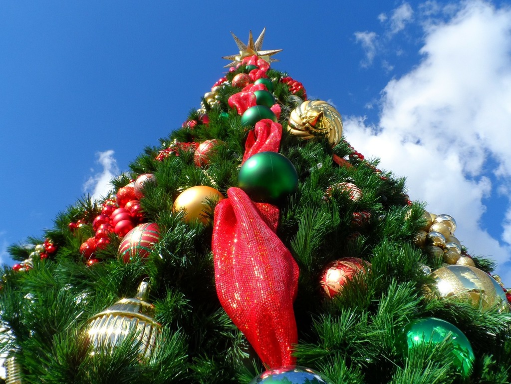 An image of a decorated Christmas tree from the ground looking up into a blue sky with partial clouds. The tree is green with green, gold, red, and silver bulbs, with a 10 point star on top.