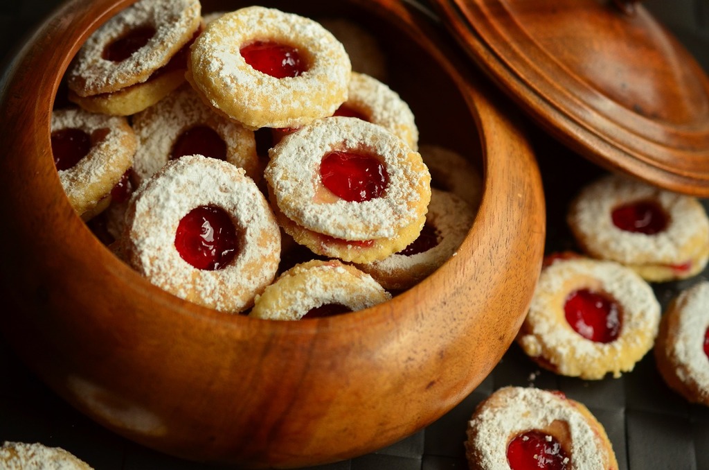 An image of a wooden bowl, with the lid propped off to the upper right hand side, and inside the bowl are jelly cookies with red jelly on the inside, shortbread covered in powdered sugar on the outside. hThere are 4 and a half of these cookies also situated outside the bowl in the lower right hand corner.