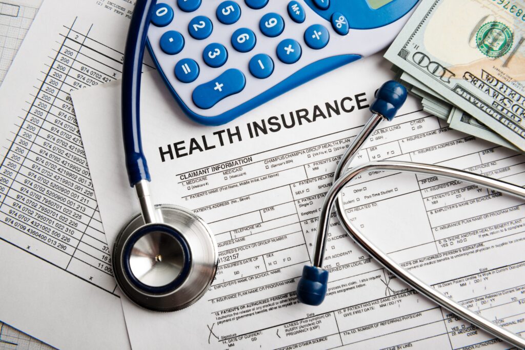 An image of a stethoscope, calculator, a small pile of 100 dollar bills sitting on top of an incomplete health insurance form.