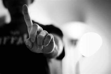 A black and white image of a person, blurred in the background, holding up their index finger signaling in a gesture indicating to wait just a moment. 